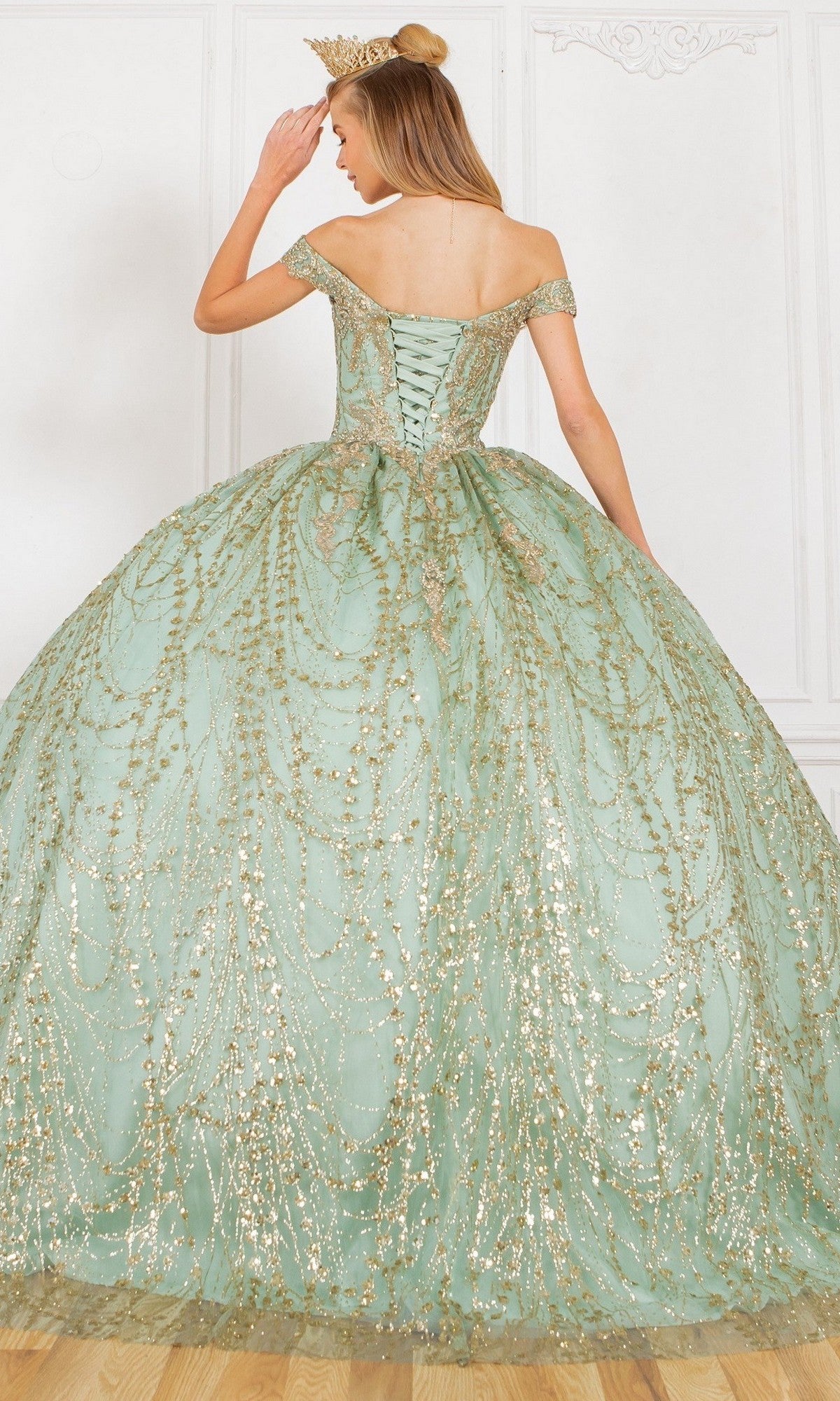 A woman wearing the Quinceanera Dress 4201 by Cinderella Couture—a pale green ball gown with gold embellishments and a crown—stands on a wooden floor, elegantly facing away in a refined room.